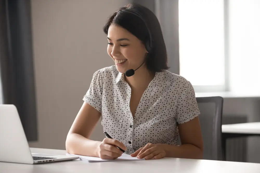 Happy asian business woman wearing headset with microphone, watching educational webinar, writing down notes. Smiling female employee working in online support service, helping client with problem.
