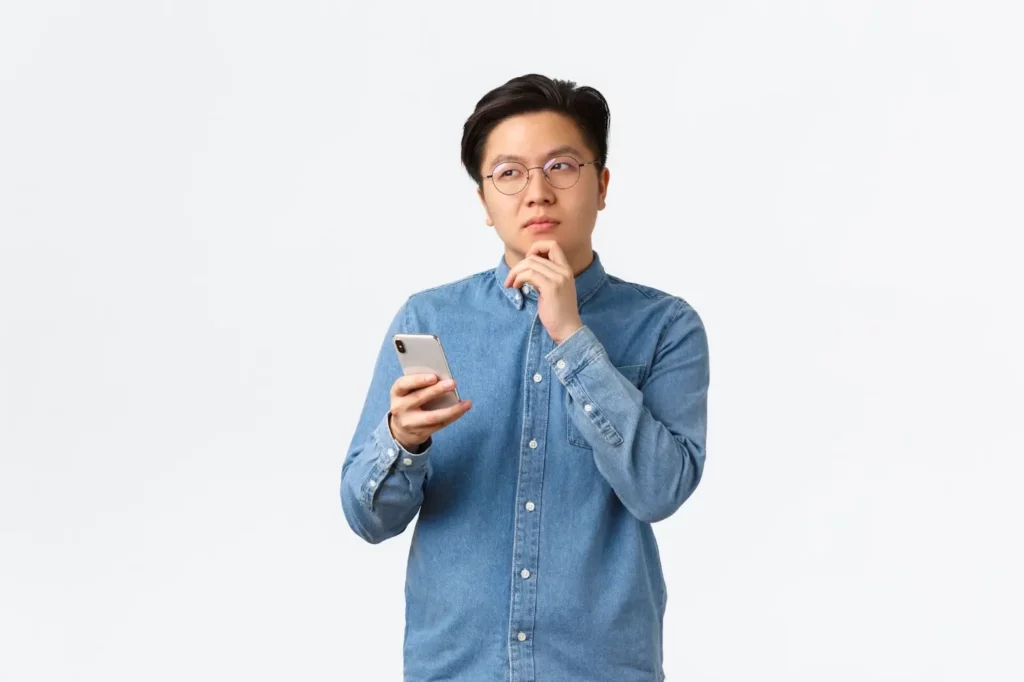 asian man in glasses holding phone and thinking with light plain background