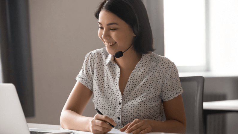 Happy asian business woman wearing headset with microphone, watching educational webinar, writing down notes. Smiling female employee working in online support service, helping client with problem.