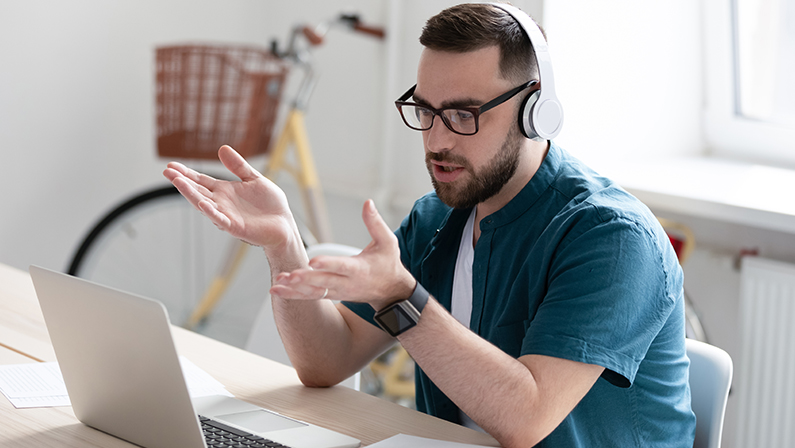 Focused young businessman in eyewear wearing headphones, holding video call with clients on laptop. Concentrated millennial man in glasses giving online educational class lecture, consulting customer.