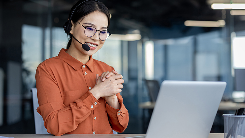 Friendly Asian businesswoman with headset at her desk in a modern office setting, exuding professionalism and warmth.