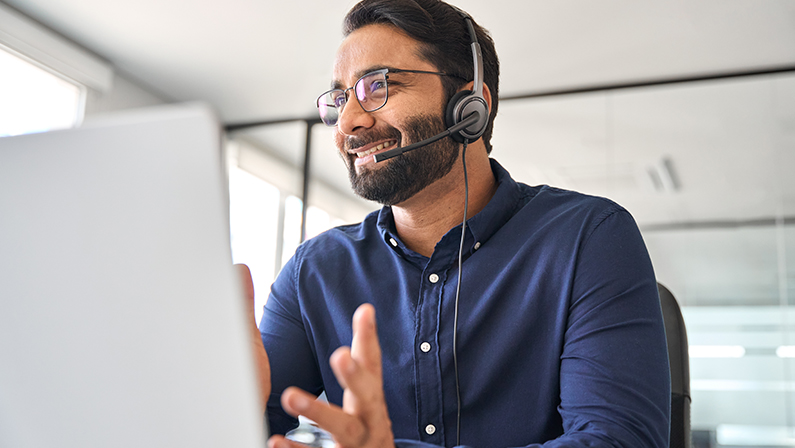 Happy Indian call center agent wearing headset talking to client working in customer support office. Professional contract service telemarketing operator using laptop having conversation. Candid shot