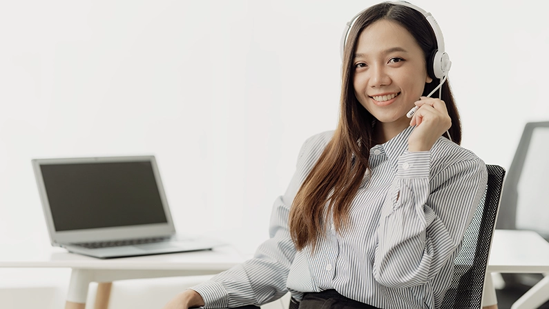 Beautiful Asian female salesperson or call center operator wearing a wireless headset is talking with a customer on the phone. Provide advice and communicate with customers via computer video calls.