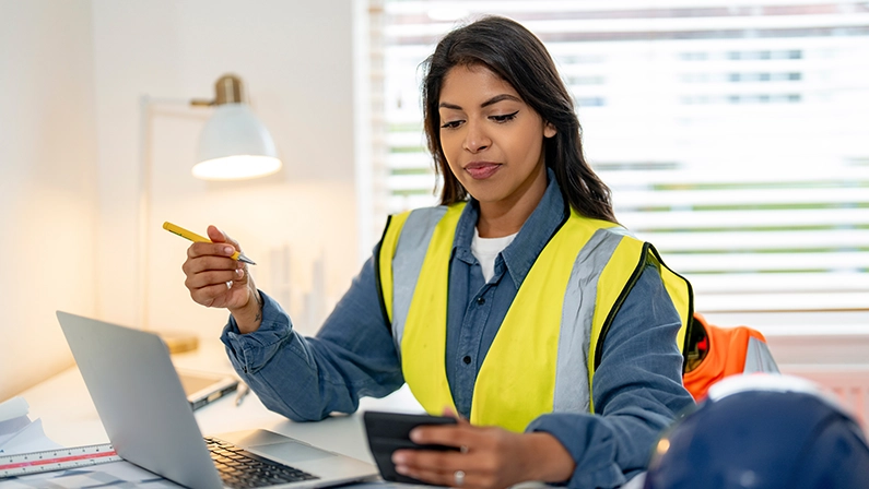 A female engineer in a high-visibility vest using a smartphone and laptop for remote construction management in an office setting.