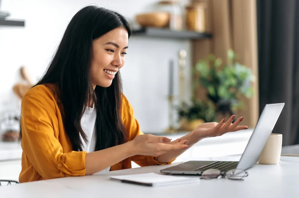 Friendly pretty Asian brunette girl, manager, freelancer, sit in the kitchen, uses laptop