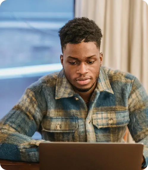 A man in a plaid shirt sits at a table, working on a laptop.