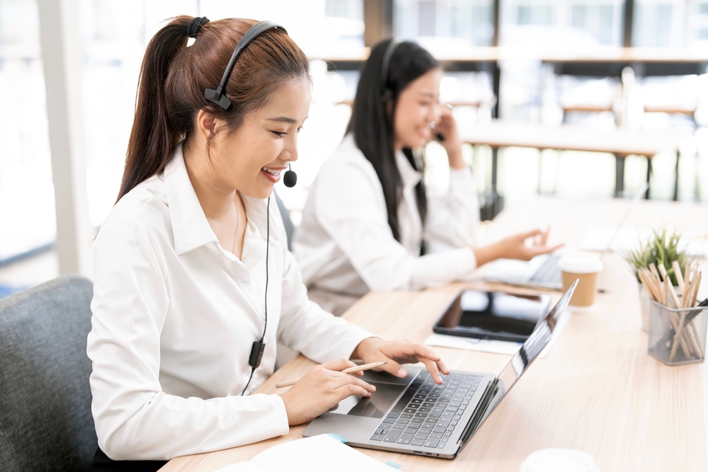 Close-up of a happy Asian call center agent wearing a headset, typing on her laptop while providing professional customer support