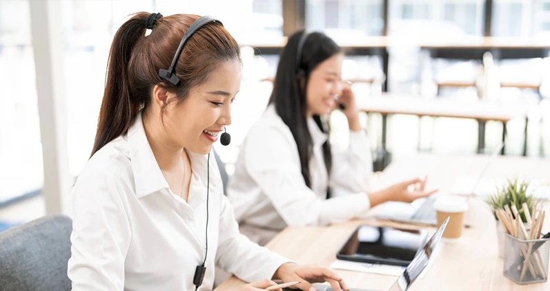 Smiling Asian woman wearing a headset with a microphone, providing excellent customer support or attending a virtual meeting at her laptop in a modern call center or office.