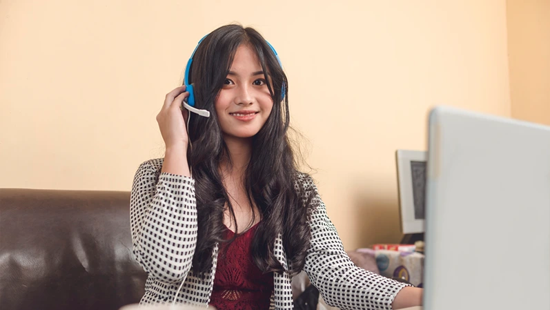 A young and attractive Filipina online teacher working at her living room. Wearing a blue wired headset.