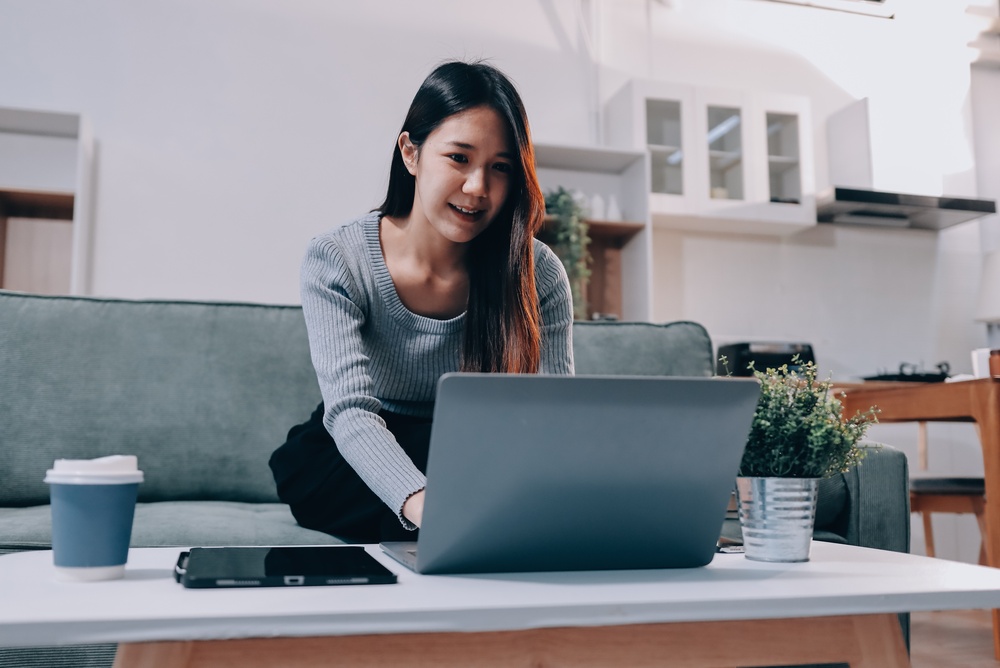 Freelance Asian woman holding mobile phone typing on laptop and working from home. Happy young girl sitting on sofa in living room.