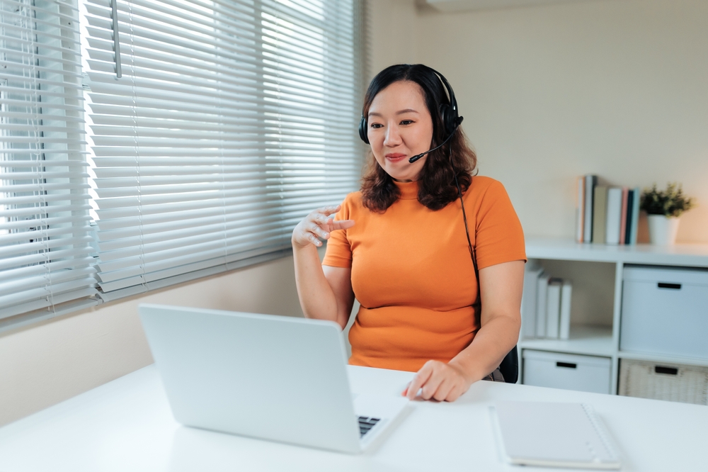 Woman wearing headset communicating during online meeting, teleworking remotely from home office using laptop